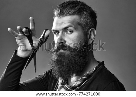 young handsome bearded man with long beard moustache and brunette hair holding hairdresser or barber scissors with emotional face in studio on grey background