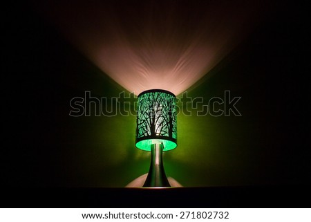 Green lamp with a tree design lighting up a wall at night surrounded by dark shadows.