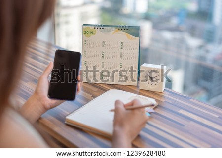 Woman holding smartphone to update calendar 2019 with notebook pencil diary on table with blurred background. Planning scheduling agenda event appointment for year 2019. Calendar and Planning concept.