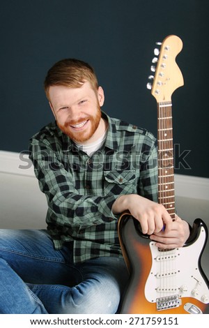 Happy young red hair man with red beard in plaid shirt holding a guitar on dark background