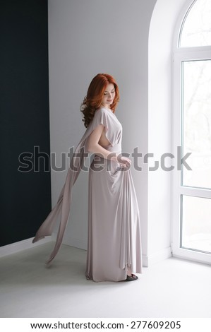 Portrait of a smiling beautiful red-haired (ginger) girl posing in studio. Looking down. Healthy long and wavy hair. Daylight.