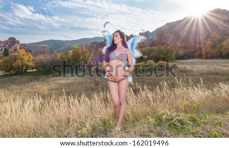 Beautiful expecting mother to be pregnant woman posing in front of nature and sunset wearing lingerie and angel wings showing belly