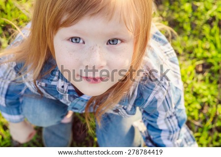 cute smiling redhead girl with freckles and blue eyes sitting on the green grass in spring Park