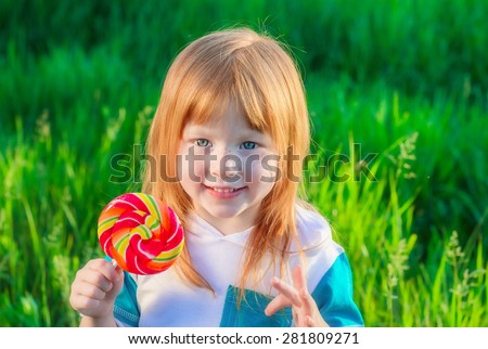 smiling red-haired girl with freckles and blue eyes holding a Lollipop on a background of green grass, soft focus