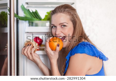 a young woman stands at the fridge and selects between an orange and a hamburger