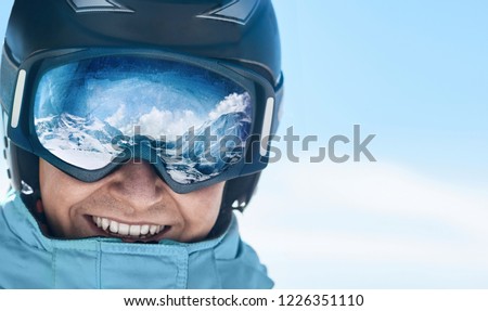 Close up of the ski goggles of a man with the reflection of snowed mountains.  A mountain range reflected in the ski mask.  Man  on the background blue sky. Wearing ski glasses. Winter Sports.