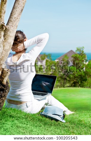 Pretty woman sitting by tree with laptop computer. Photo from behind