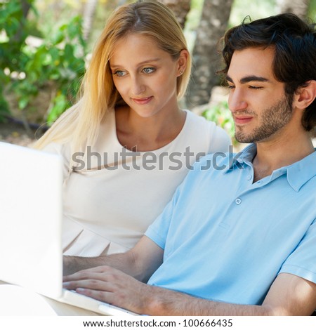 Young couple working on laptop and smiling while sitting relaxed on bench at summer park