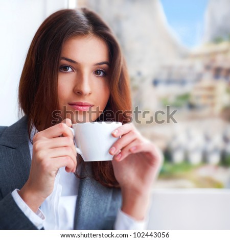 Portrait of beautiful young business woman wearing formal clothes drinking coffee while resting at her office near big window