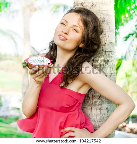 Portrait of young pretty woman wearing bright pink dress eating exotic asian dragon fruit and enjoying her vacation at tropical resort