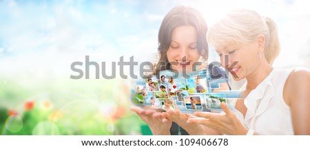 Two women sitting at park and using virtual interface to watch video or pictures