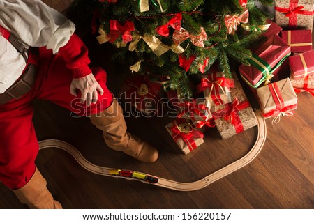 Unrecognizable Santa Claus Playing With Wooden Railroad Near Christmas Tree. Top View Indoors At Dark Room