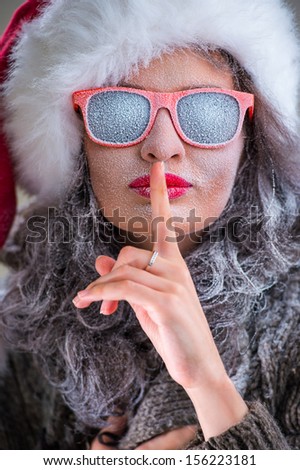 Woman wearing Santa Claus hat and sunglasses with snow on her face showing silence gesture. Fashion portrait