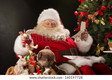 Santa Claus sitting at home at comfortable armchair holding envelope and reading children\'s letters and wishes and choosing toys from big sack near him.