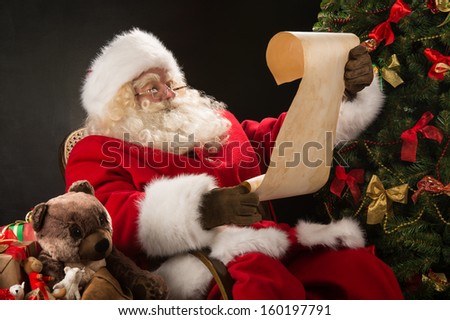 Portrait Of Happy Santa Claus Sitting At His Room At Home Near Christmas Tree And Big Sack And Reading Christmas Letter Or Wish List