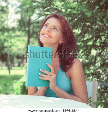Positive adult beautiful woman sitting near table and reading book outdoors at backyard in summer. Retro retouch photo