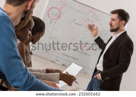 Business meeting at office. Handsome man presenting charts on whiteboard to team. Multi ethnic group of people
