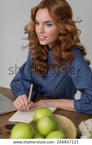 Happy calm woman writing something in diary or notepad at her workplace at home or office, flowers and green apples on table