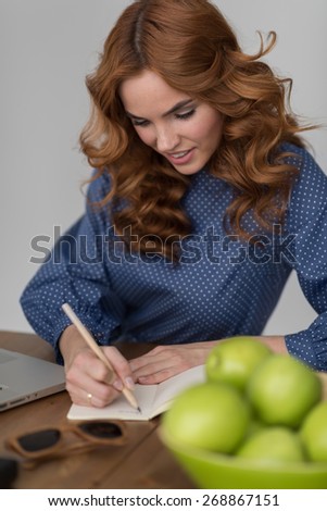 Happy calm woman writing something in diary or notepad at her workplace at home or office, flowers and green apples on table