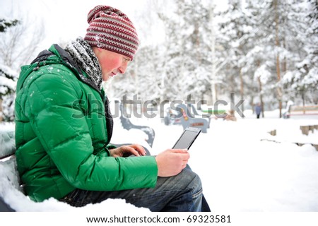 Portrait of young man outdoor in winter park wearing bright clothes working with his netbook and enjoying free wireless internet connection