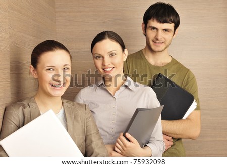 Group of business associates standing together against modern wooden wall at their office one woman holding laptop, man and other woman holding folder with papers and documents. Young entrepreneurs.