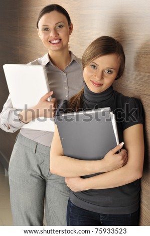 Portrait of two young handsome women standing relaxed against modern wooden wall in their office smiling and holding laptop and folder with papers