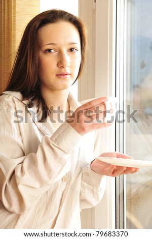 Portrait of young girl standing near a window at home. Drinking coffee and warming in sunbeam