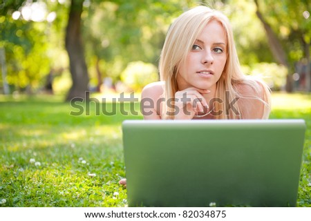 Portrait of young pretty woman resting on green grass at summer park using laptop to communicate her friends. Outdoors. Daydreaming