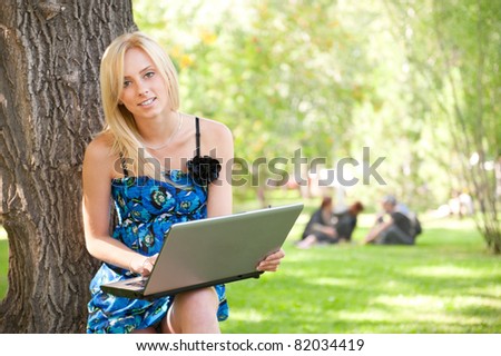Portrait of a pretty student woman leaning on the tree trunk on foreground at summer park. Group of students at the background