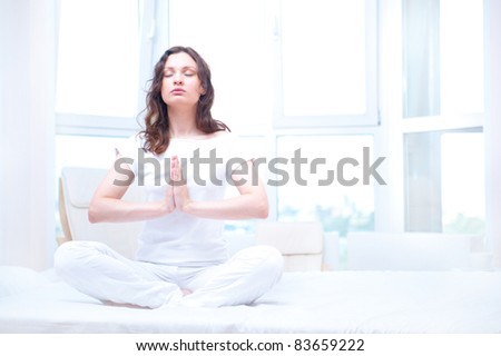Young woman meditating with closed eyes in bright bedroom sitting on bed