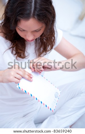 American or european young pretty woman sitting on bed in bright bedroom and opening letter. She is excited and dreaming about enrollment to her favorite college or university.