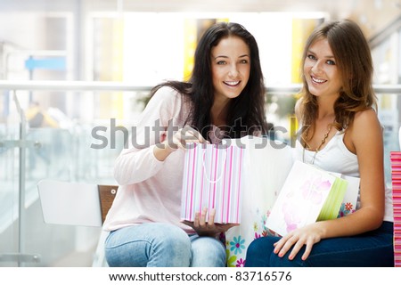 Two excited shopping woman resting on bench at shopping mall looking at camera