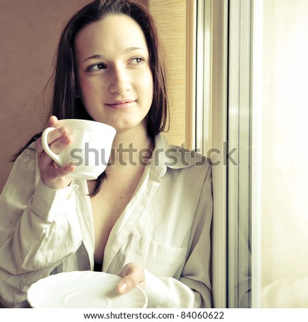 Portrait of cosy young girl standing near a window at home. Drinking coffee and warming in sunbeam