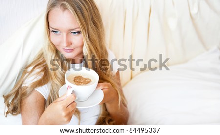 Smiling woman drinking a coffee lying on a bed at home or hotel. Heart shape illustrated on coffee foam. Horizontal shot. Model is looking away. Lots of Copyspace.