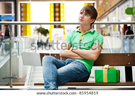 Portrait of young man inside shopping mall with gift box sitting relaxed on bench and waiting for his girlfriend