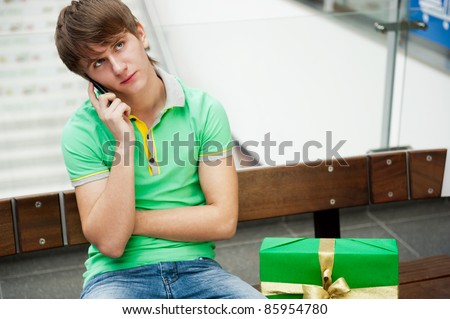 Portrait of young man inside shopping mall with gift box sitting relaxed on bench and waiting for his girlfriend