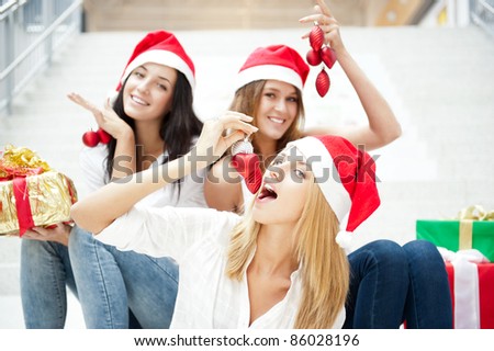 Group of three beautiful girls sitting on stairs at shopping mall hall wearing santa clause red hats with open arms