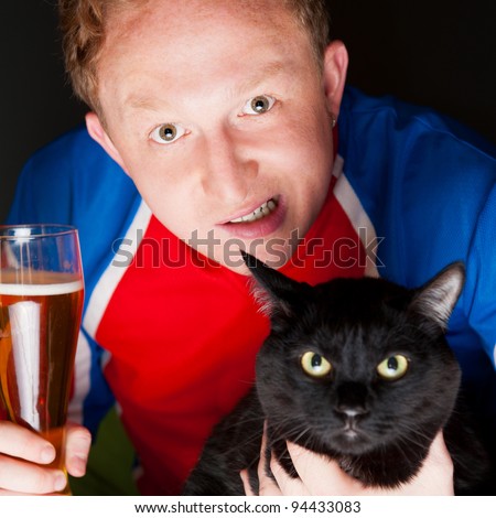 Portrait of young man holding a glass of beer and a big black cat and both looking at camera while watching tv translation of their favorite football team