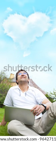 A smiling man with laptop outdoor sitting at his backyard and looking up to the cloud of heart shape overhead