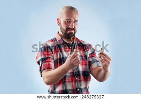 Bald and bearded guy is holding makeup brush and lipstick with real disgust expression on his face