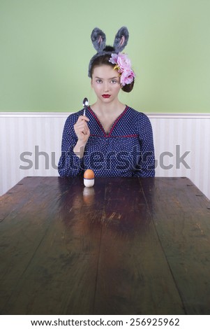 Portrait of a young woman with bunny ears sitting at the end of an empty table with an egg and spoon.