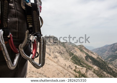 Climbing gear hanging from a hip in the foreground. Mountains, sky, and winding road in the background. Blue-grey hue.