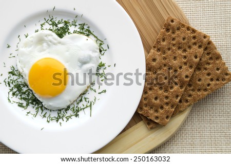Heart shape fried egg in a white plate. With cutting board and crisp bread.