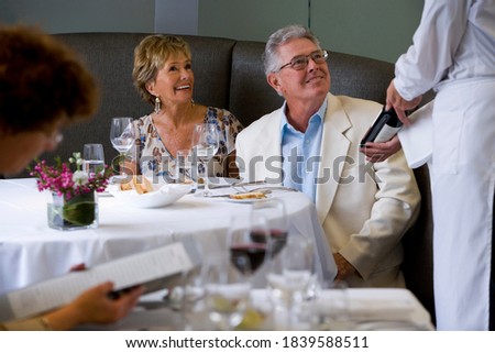 Portrait of a senior couple dining and looking at a server showcasing wine bottle in a restaurant.