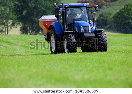Front view of a man driving a blue colored tractor in his field spreading fertilizer on a bright sunny day in a green field.