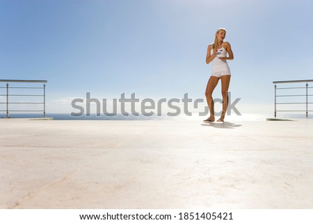 Horizontal low angle full length portrait of a happy young woman with a breakfast bowl on a deck by the sea with copy space.