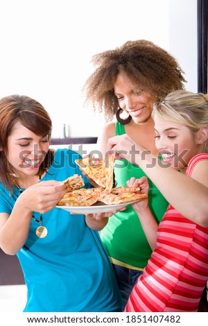 Vertical waist up portrait of three happy teenage girls picking up a pizza slice from the plate.