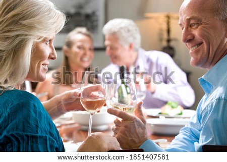 Horizontal profile shot of a mature couple toasting wine glasses at a dinner table with another couple smiling in the background.