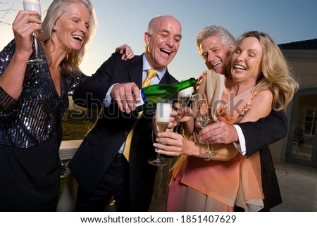 Three quarter length shot of two jubilant mature couples drinking champagne outdoors with a man pouring wine in glasses.