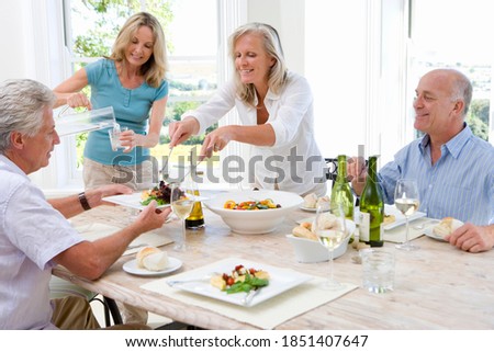 Horizontal shot of two joyous mature couples at a dinner table with the women serving drinks and food.